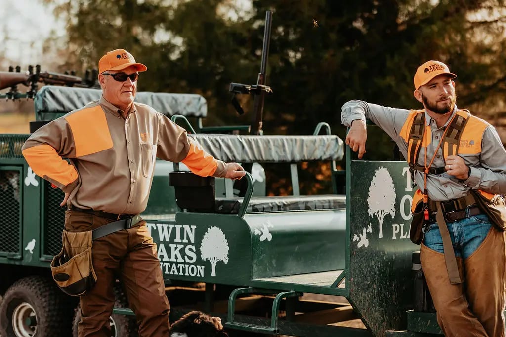 Two men in hunting attire stand next to a hunting vehicle.