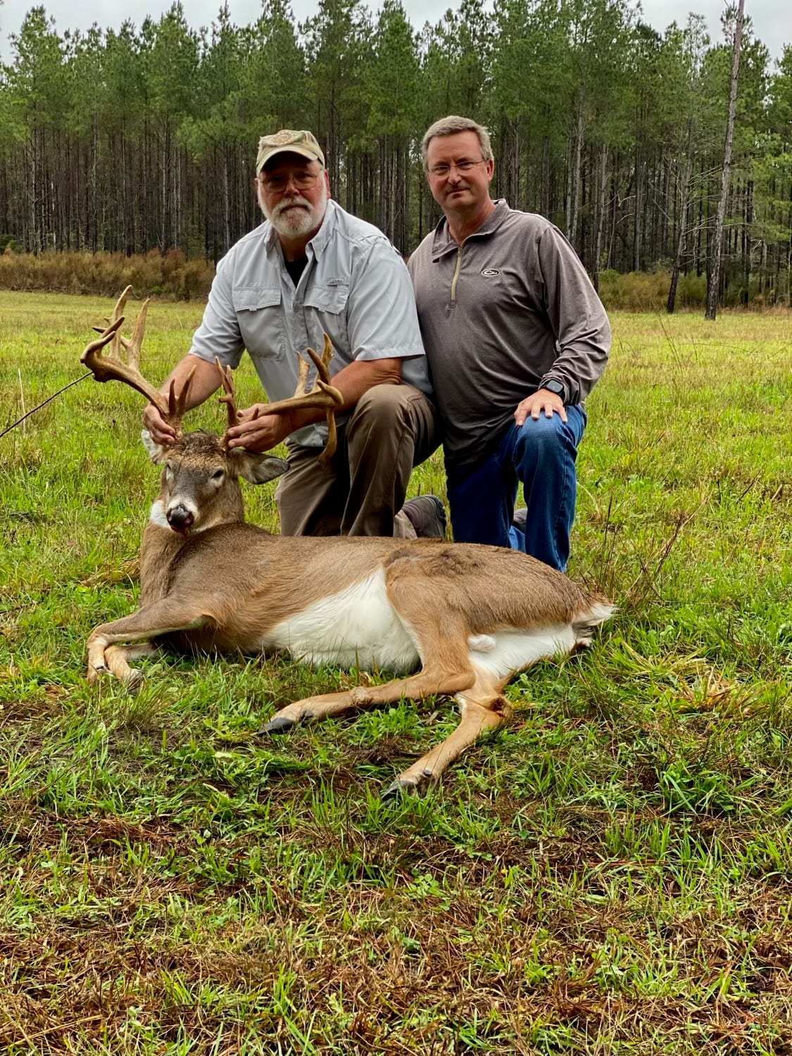 Two hunters pose with a harvested whitetail deer in a grassy field.