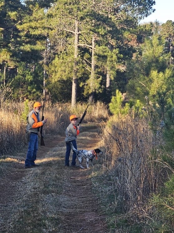 Two hunters and a hunting dog walk along a dirt path in a wooded area.