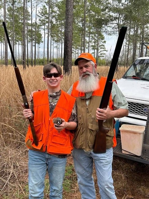 Two hunters pose with a harvested northern bobwhite quail and shotguns in a field.