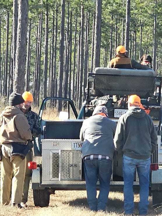 Several men in hunting attire are gathered around the back of a truck equipped with dog crates in a wooded area.
