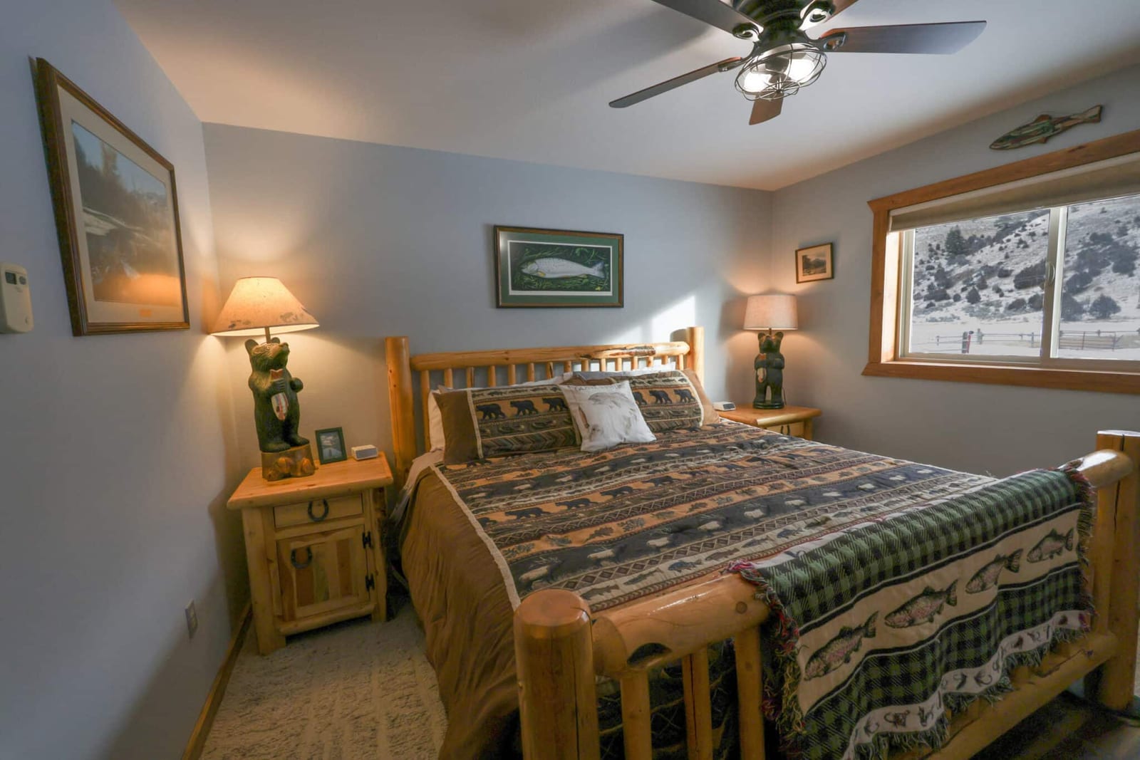 A rustic bedroom features a wooden bed frame with patterned bedding, two nightstands with lamps, and a window overlooking a snowy landscape.