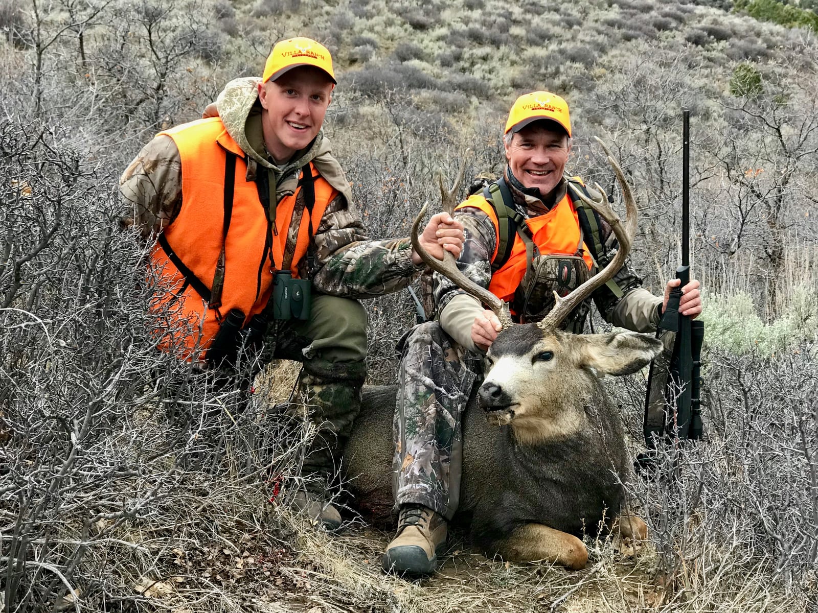 Two hunters pose with a harvested mule deer in a brushy outdoor setting.