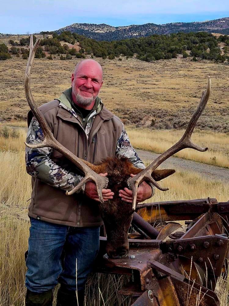 A man poses with a harvested elk in a field with hills in the background.