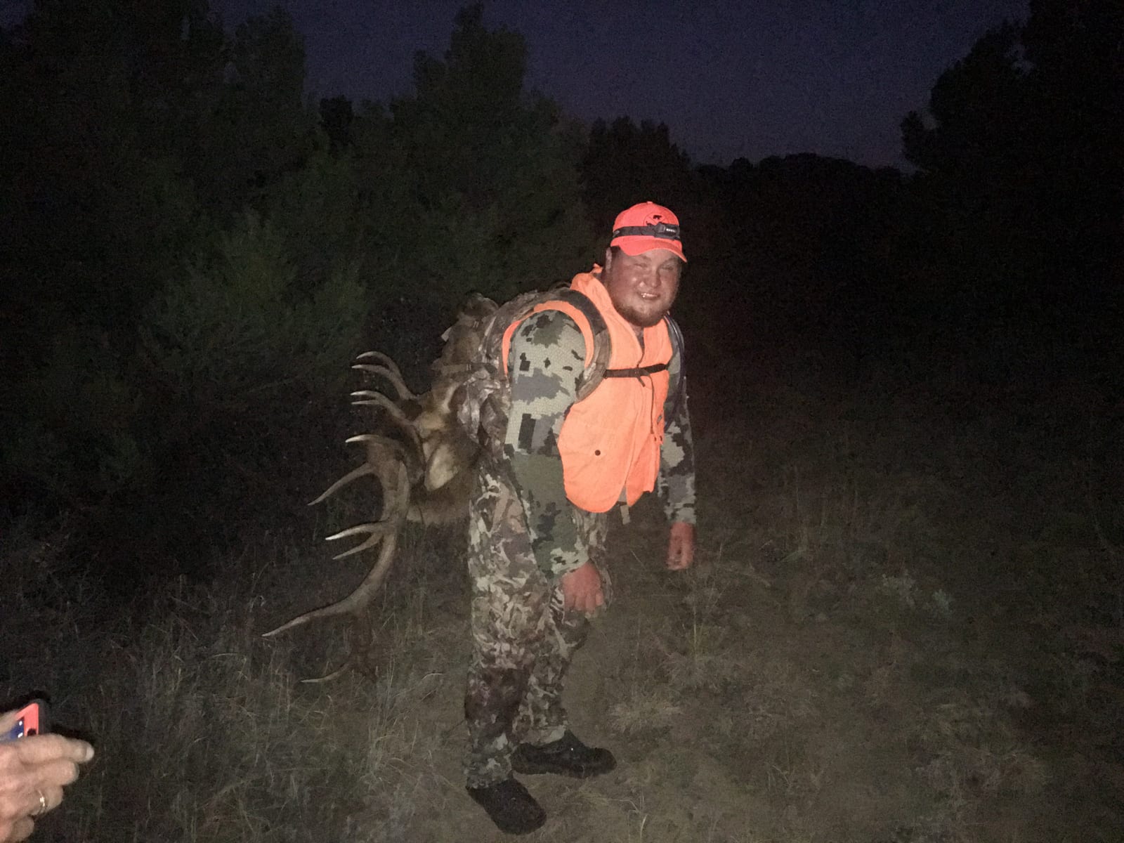 A hunter in camouflage and an orange vest carries a harvested mule deer with antlers on his back at night.
