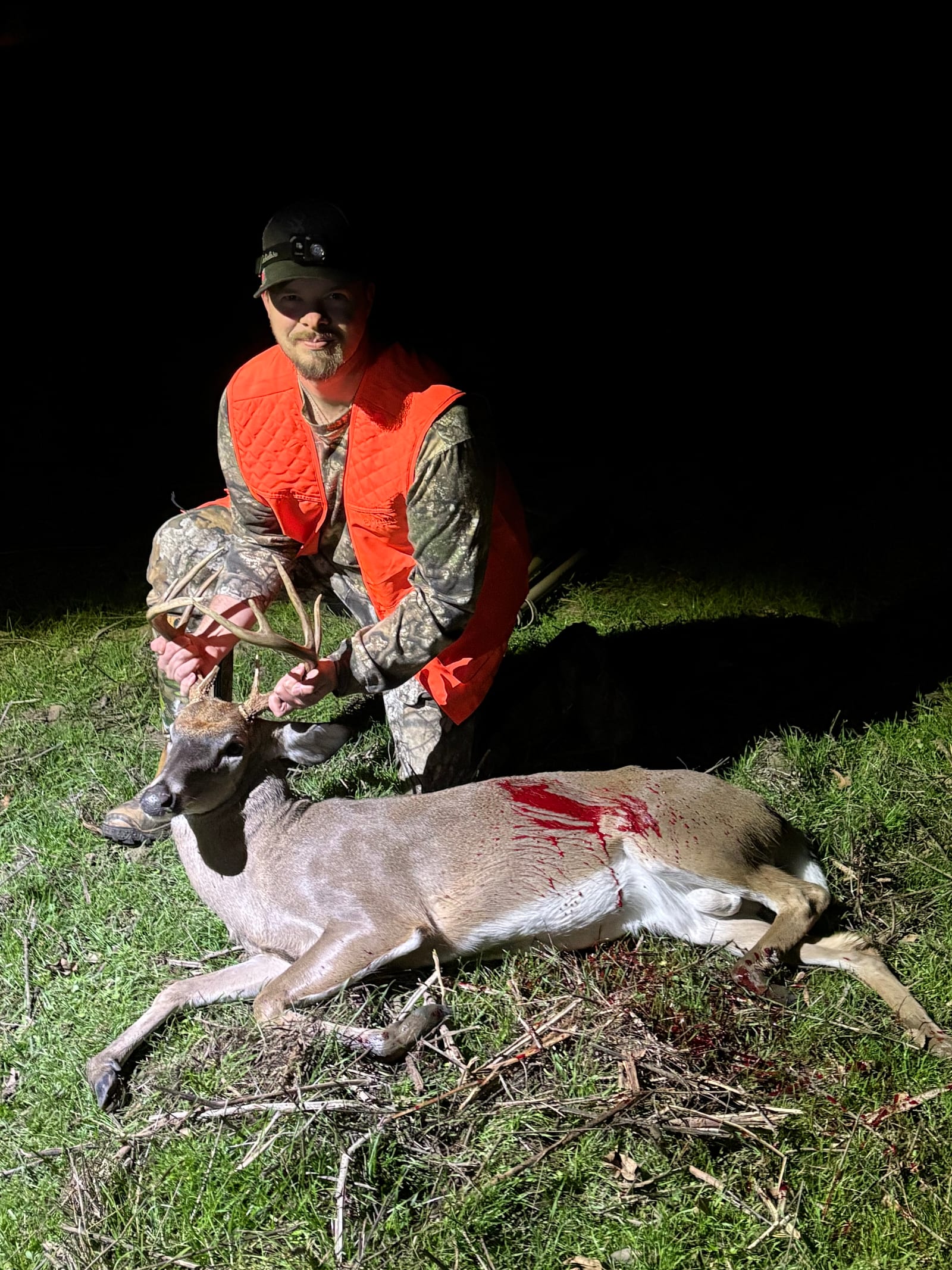 A hunter in an orange vest poses with a harvested whitetail deer at night.