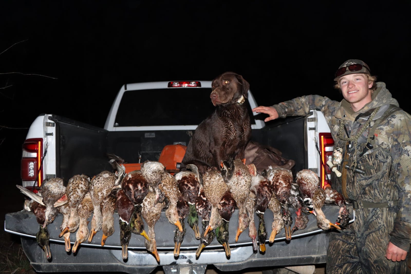 A hunter and a dog pose with a harvest of mallard and teal ducks in the back of a pickup truck at night.