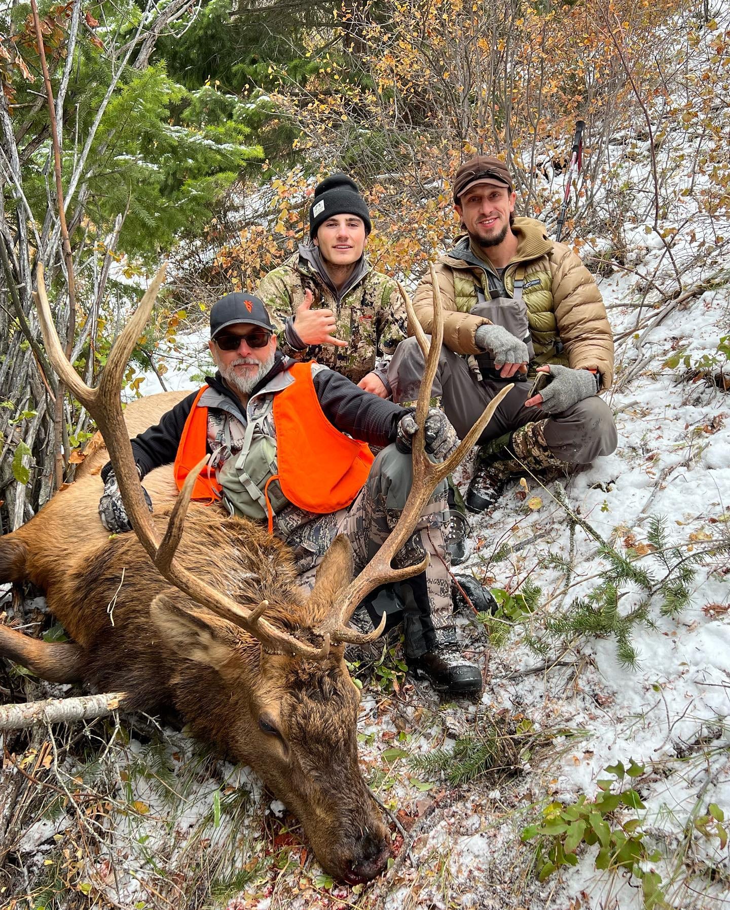Three hunters pose with a harvested elk in a snowy, wooded area.