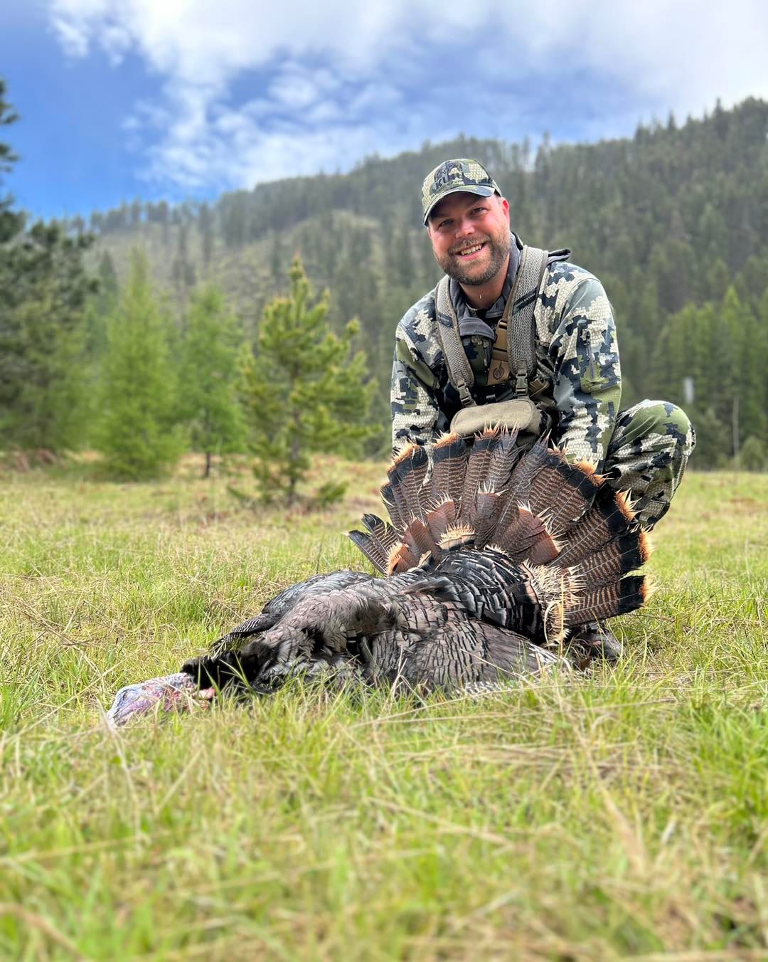 A hunter poses with a harvested wild turkey in a grassy field with trees and hills in the background.