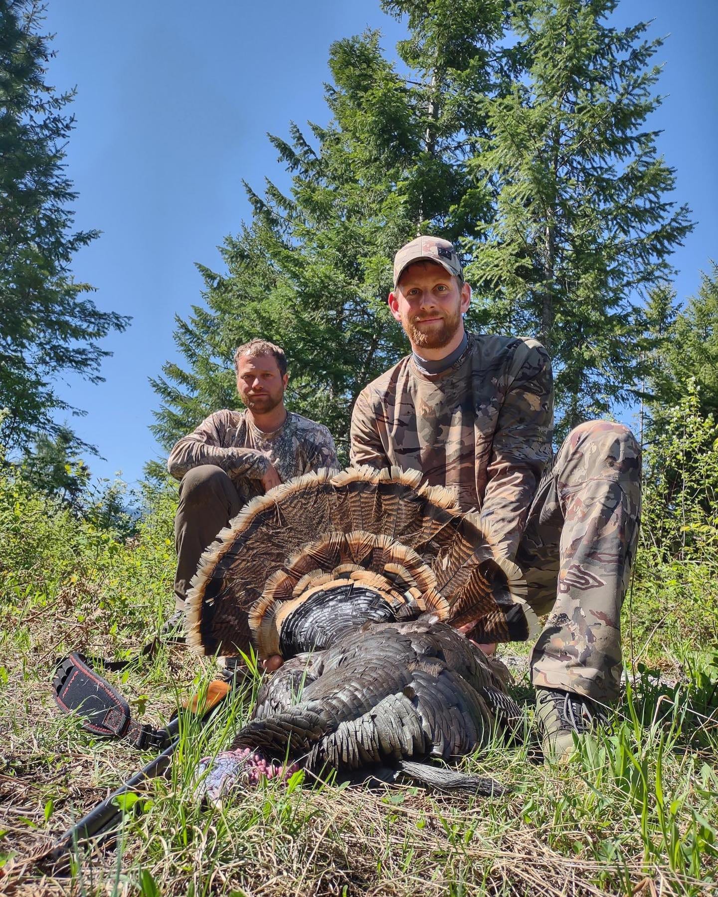 Two hunters in camouflage pose with a harvested wild turkey in a forest.
