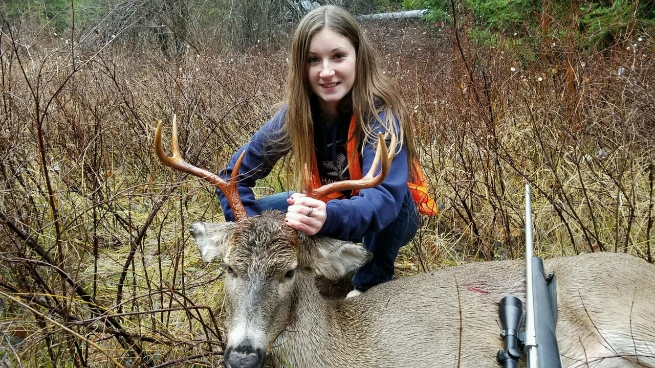 A young woman kneels next to a harvested mule deer in a brushy forest, holding its antlers, with a rifle visible on the ground.