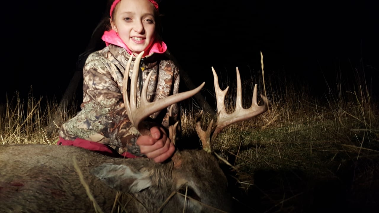 A young girl poses with a harvested whitetail deer at night.