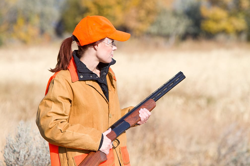 A woman in an orange hat and hunting jacket holds a shotgun in a field.