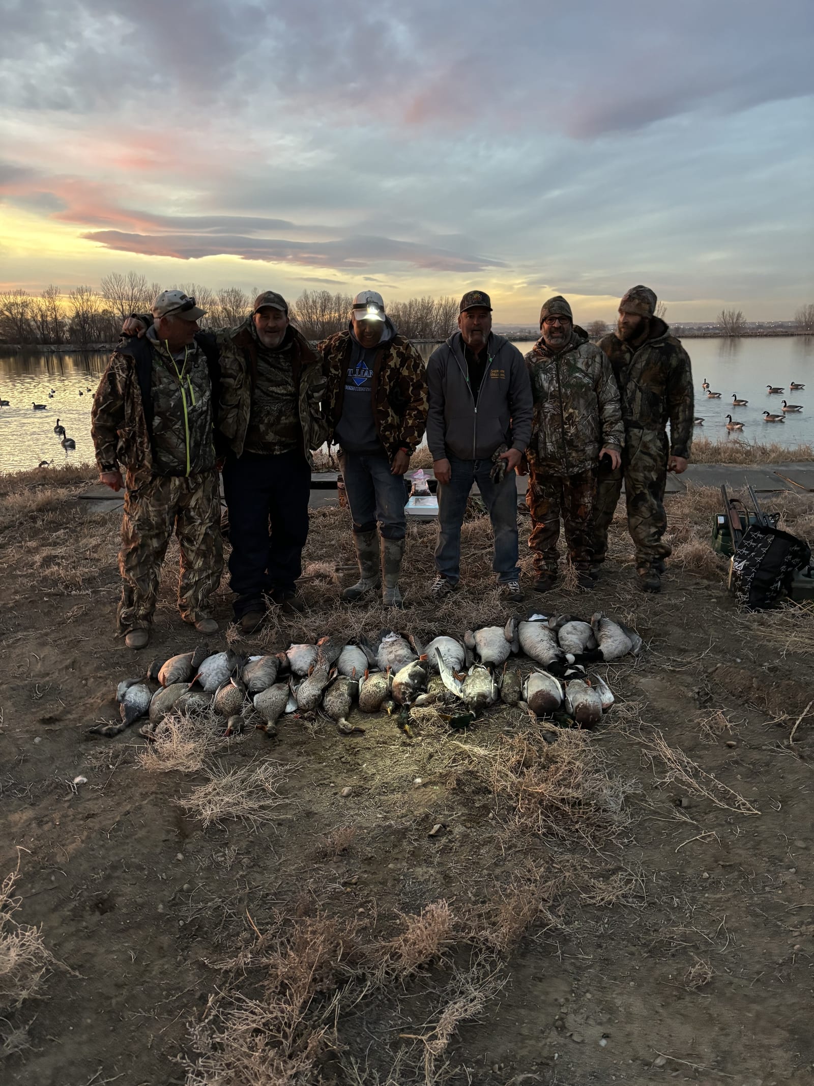 A group of six hunters poses with their waterfowl harvest, including mallards, gadwalls, american widgeons, and northern pintails, by a lake at sunset.