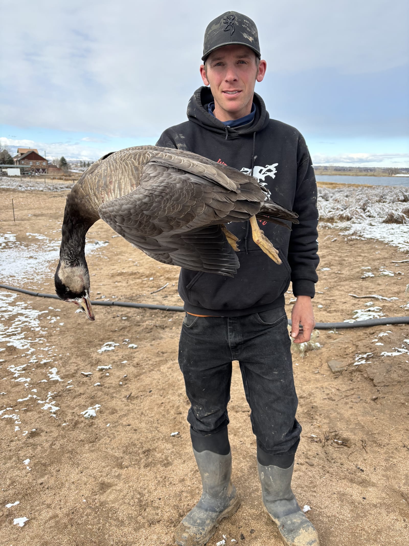 A hunter holds a harvested Canada goose in a field with a snowy, muddy ground and a body of water in the background.