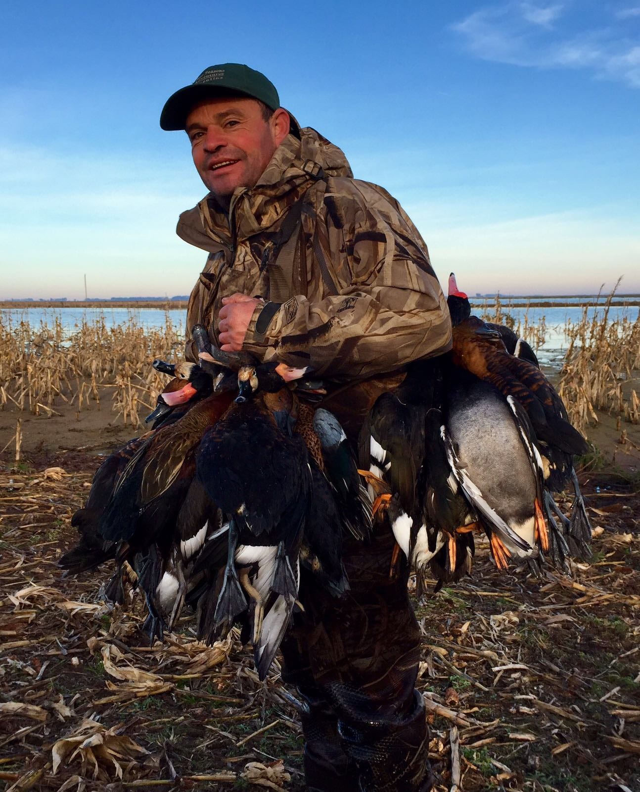 A hunter in camouflage holds a string of harvested ducks or geese in a field near water.