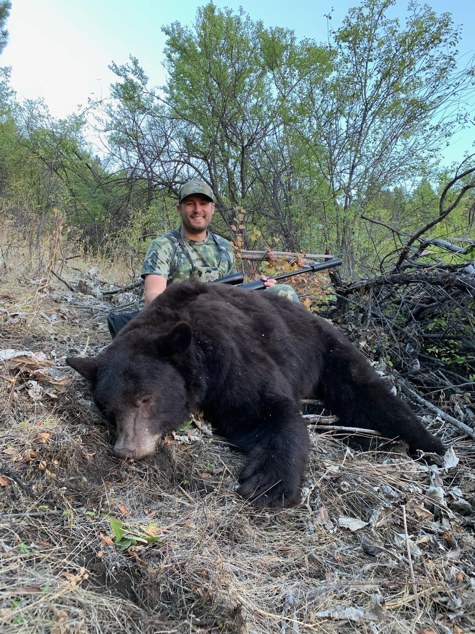 A hunter in camouflage poses with a harvested black bear in a wooded area.