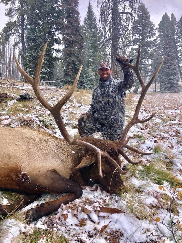 A hunter poses with a harvested elk in a snowy, forested area.