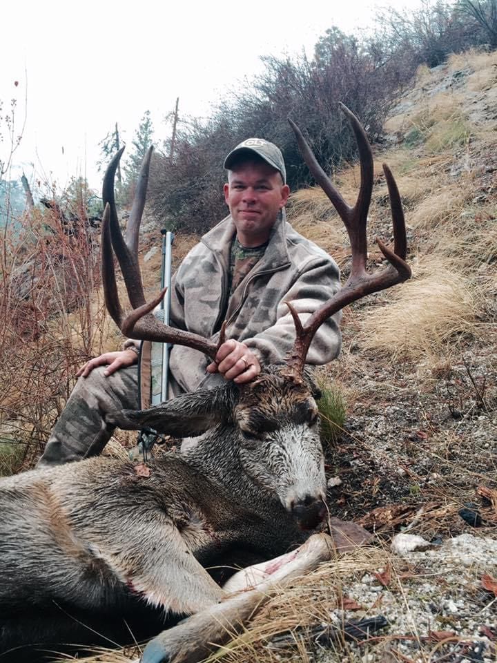 A hunter in camouflage clothing poses with a harvested mule deer on a hillside.