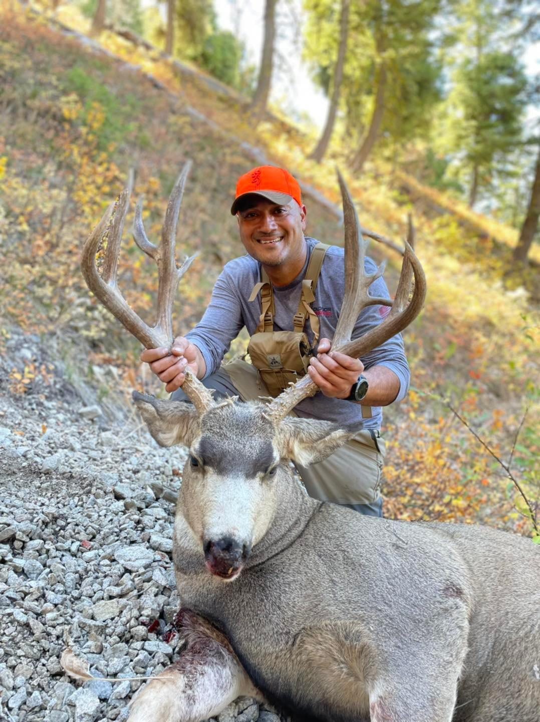 A hunter in an orange hat poses with a harvested mule deer on a rocky hillside.