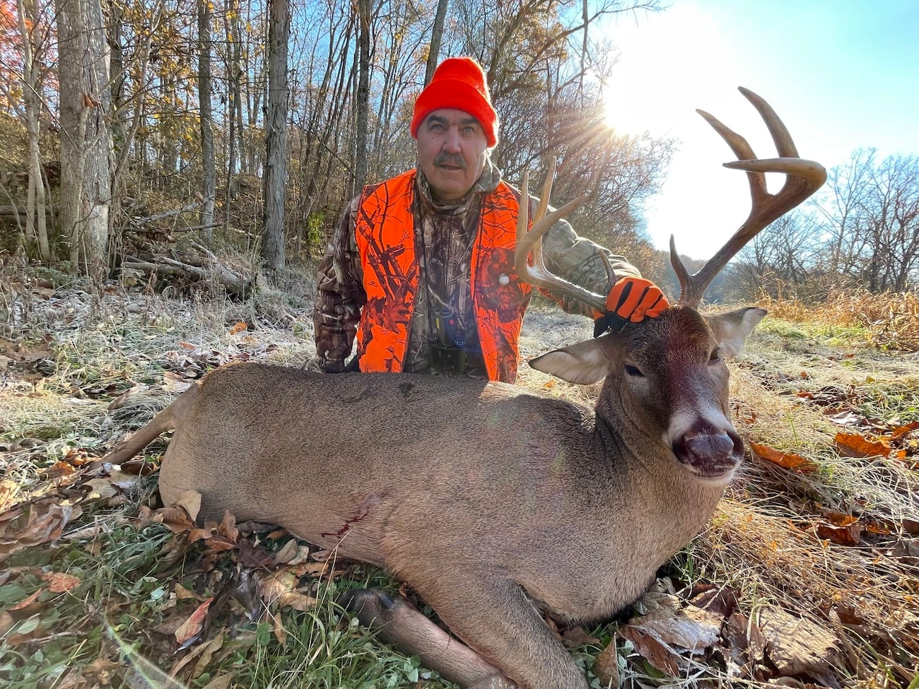 A hunter in an orange hat and vest poses with a harvested whitetail deer in a wooded area.