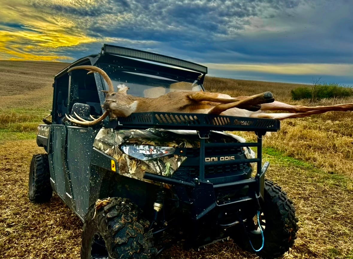 A harvested whitetail deer is loaded onto the back of a UTV in a field under a dramatic sky.