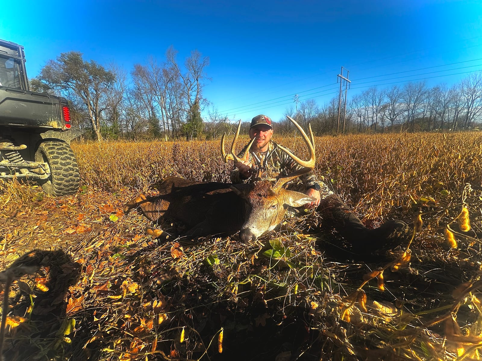 A hunter poses with a harvested whitetail deer in a field next to an ATV.