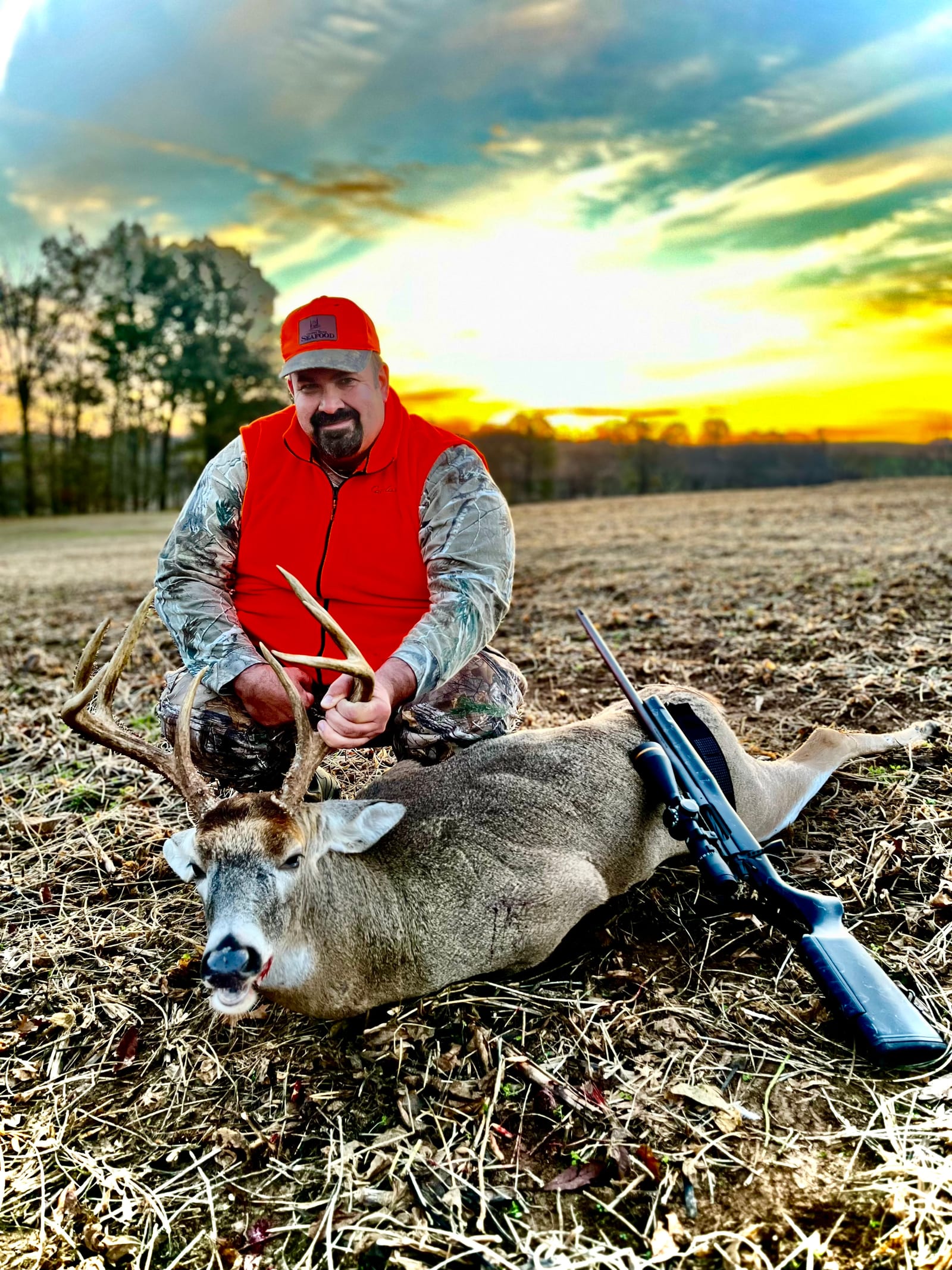 A hunter kneels beside a harvested whitetail deer in a field at sunset.