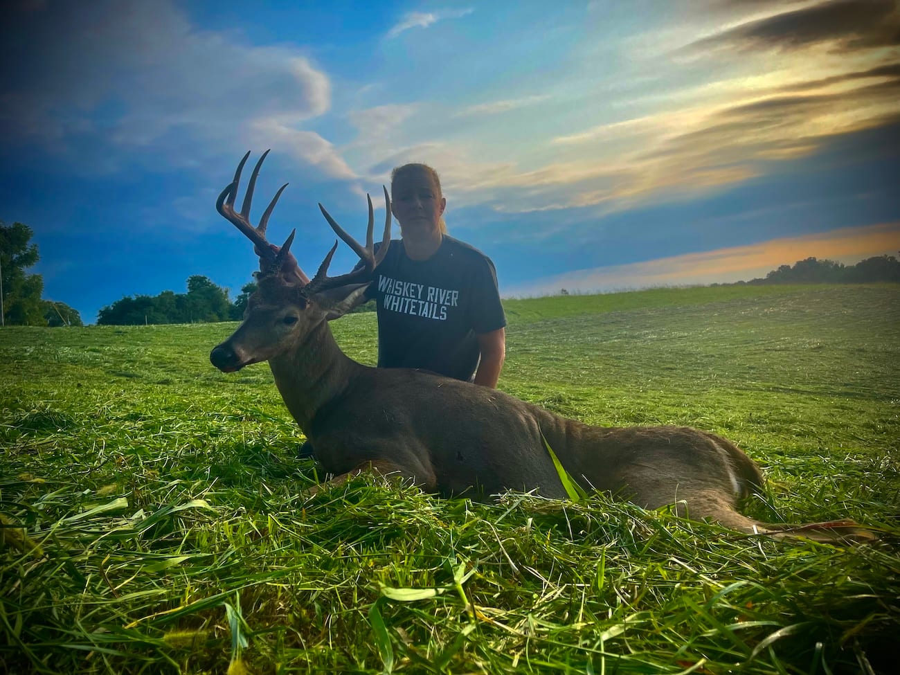 A hunter poses with a harvested whitetail deer in a grassy field at sunset.