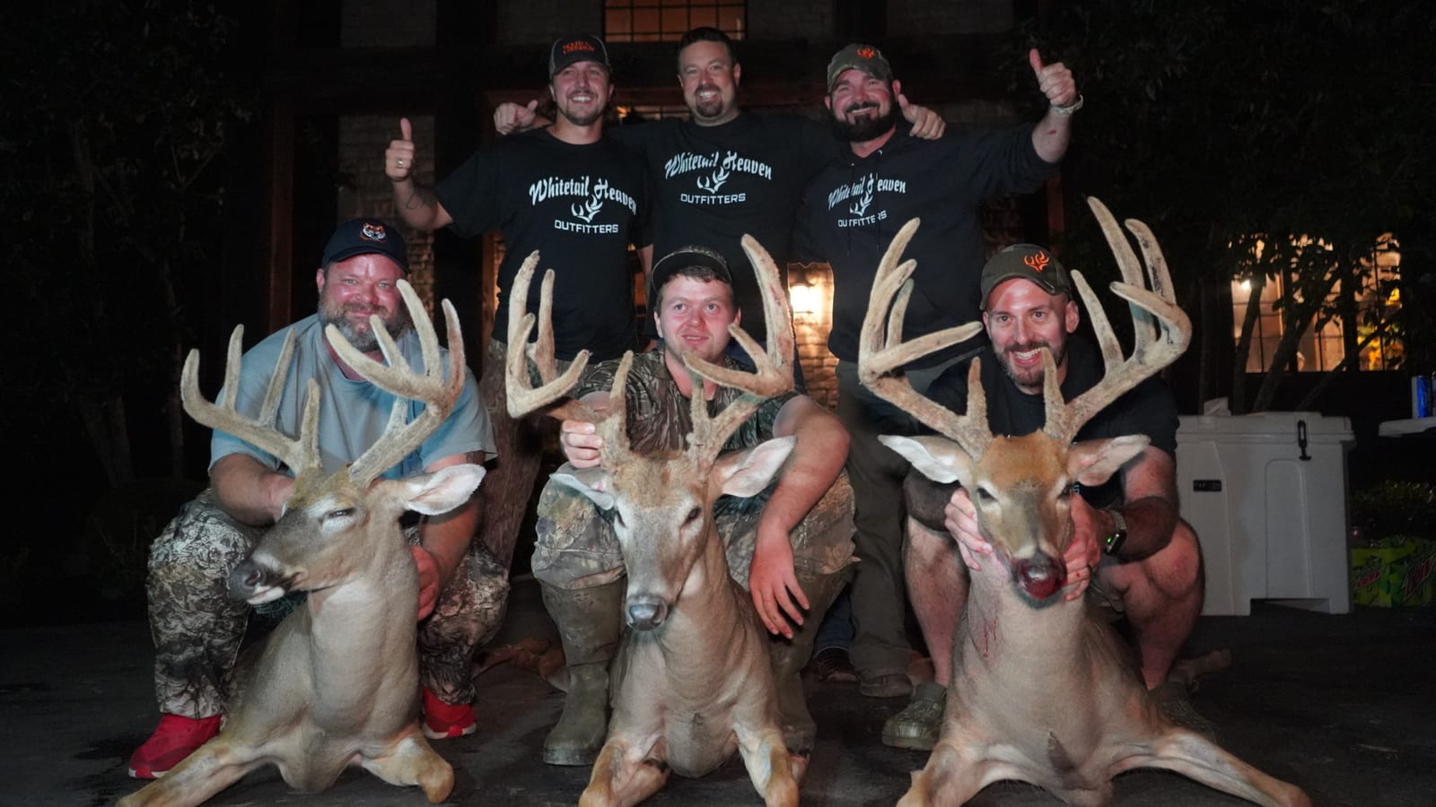 Several men pose with three harvested whitetail deer at night.