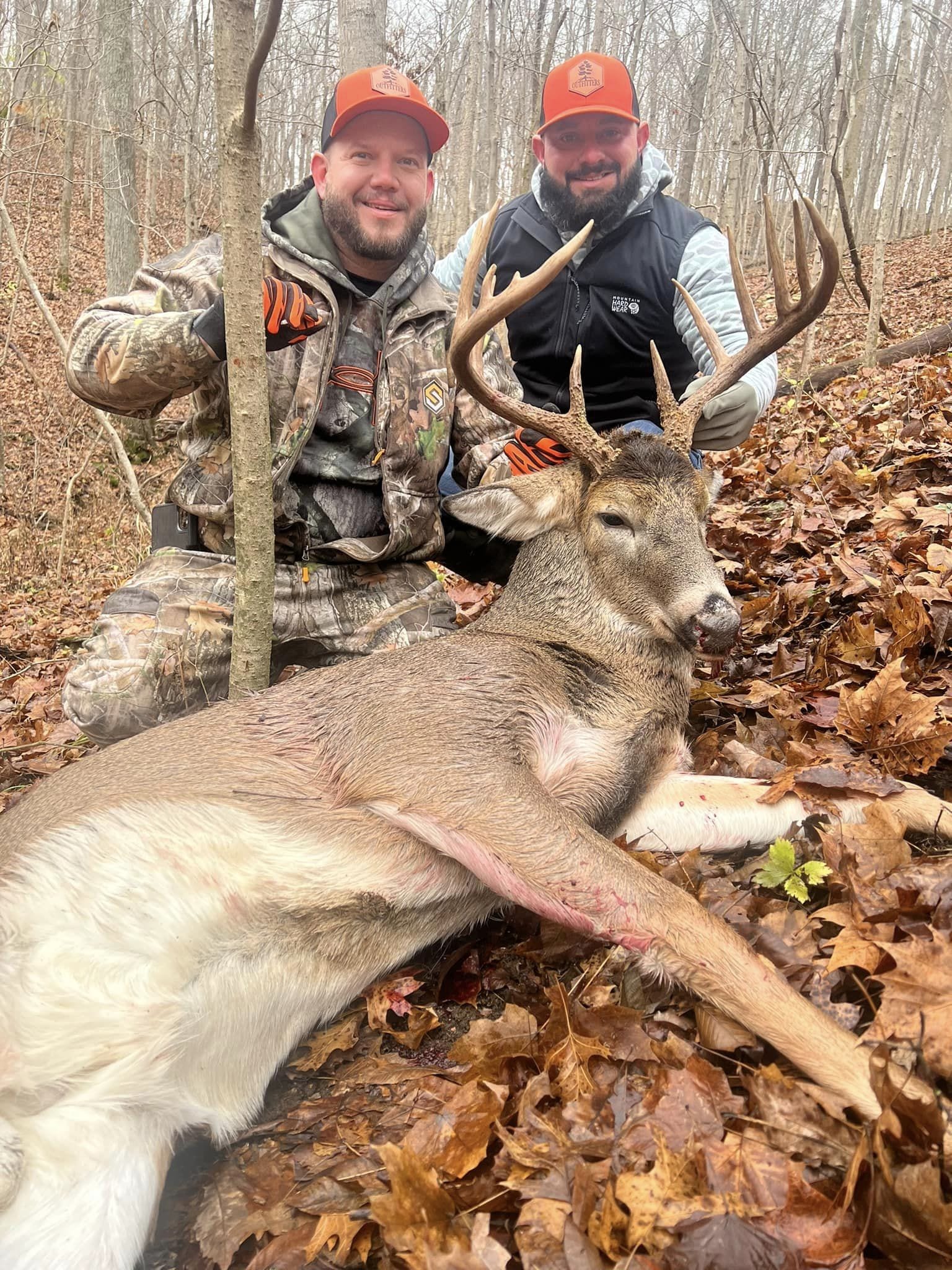 Two hunters pose with a buck on a leaf‑covered forest floor.