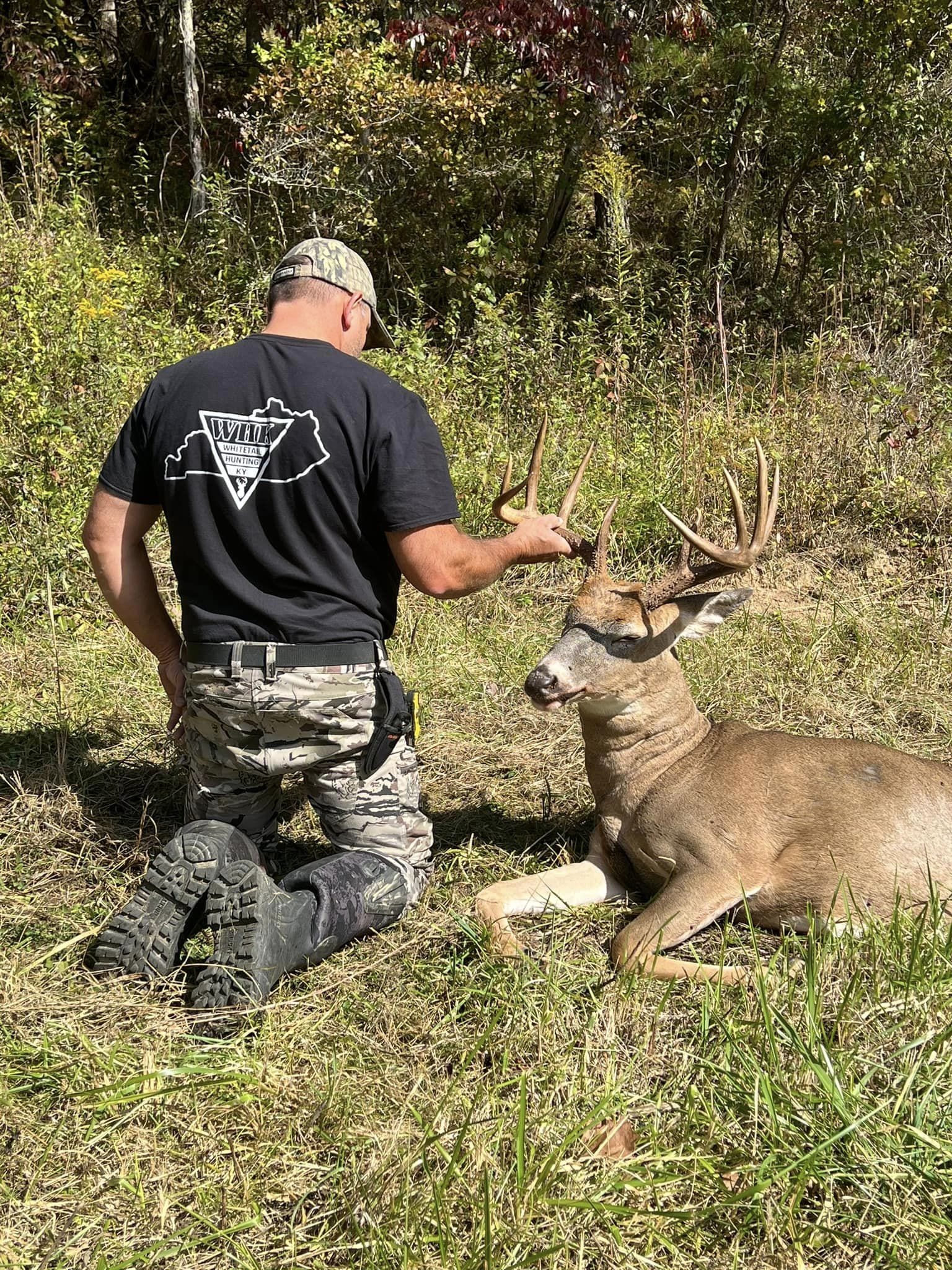 A hunter kneels beside a large buck in a grassy field with autumn foliage in the background.