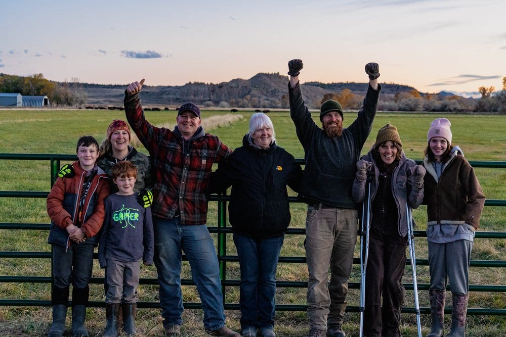 A group of adults and children pose in a field with mountains in the background.
