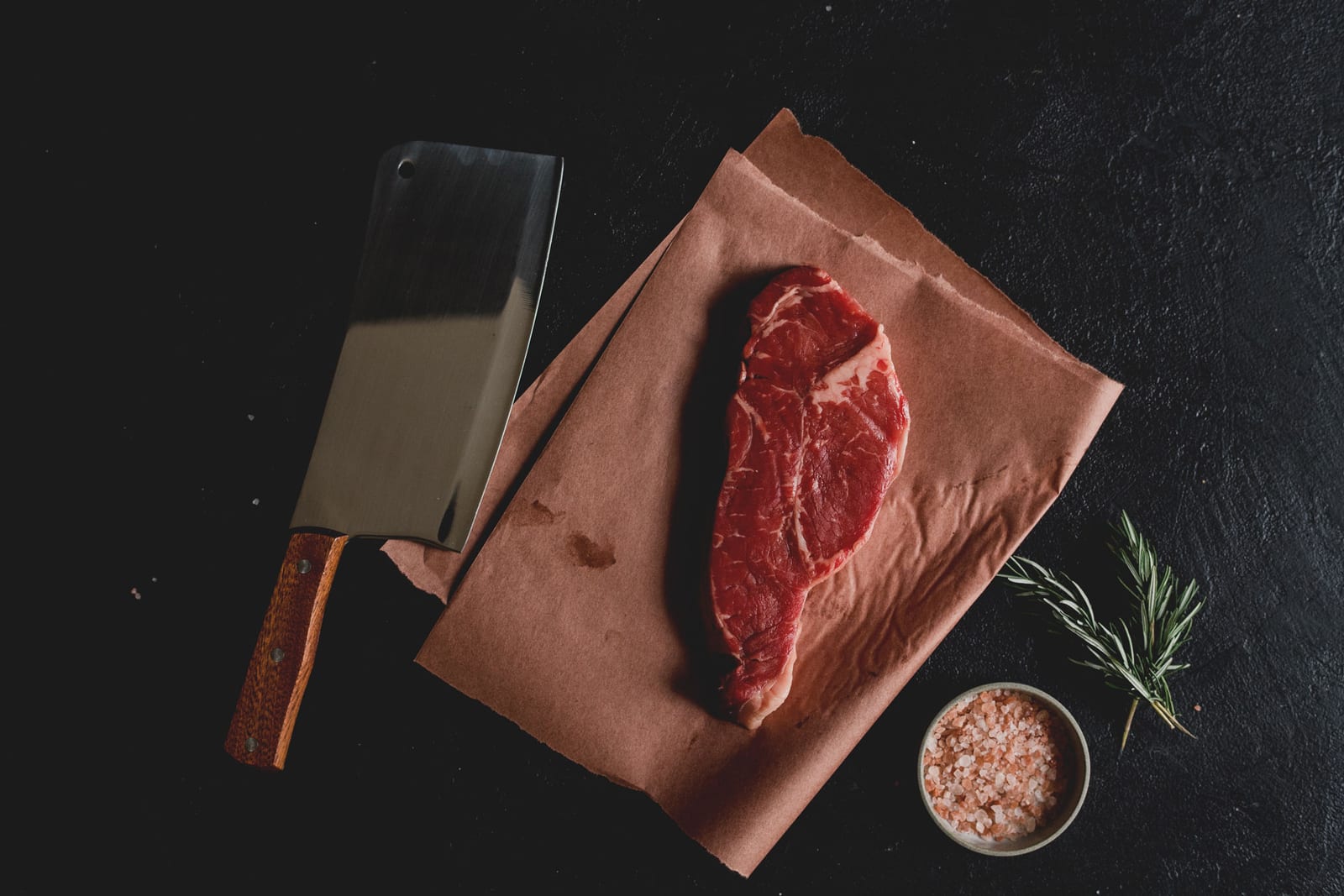 A raw steak on brown paper with a cleaver, salt, and rosemary on a dark surface.