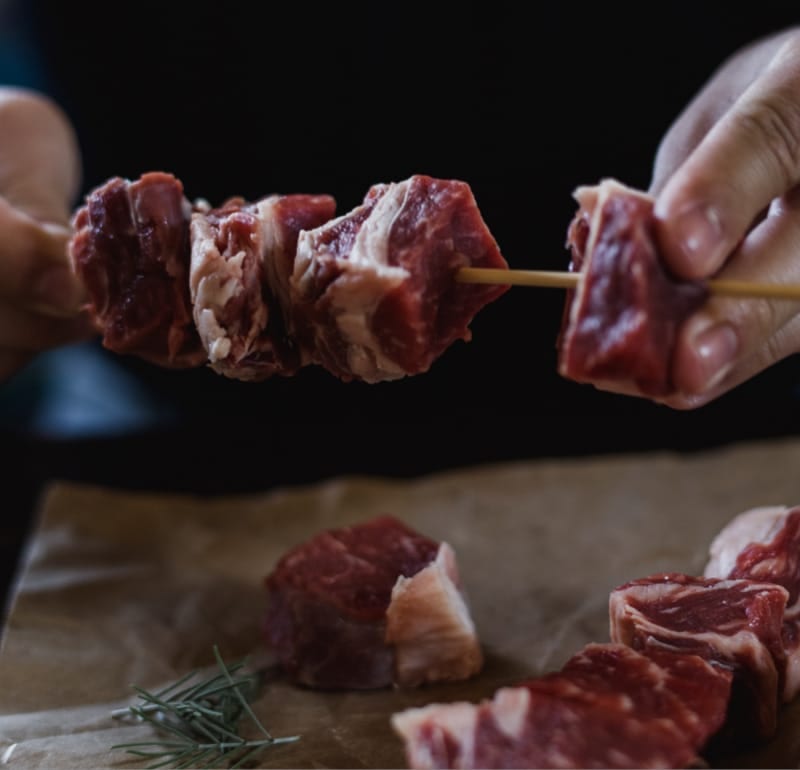 Hands are skewering raw meat onto a wooden stick, with more raw meat and rosemary sprigs on brown paper below.