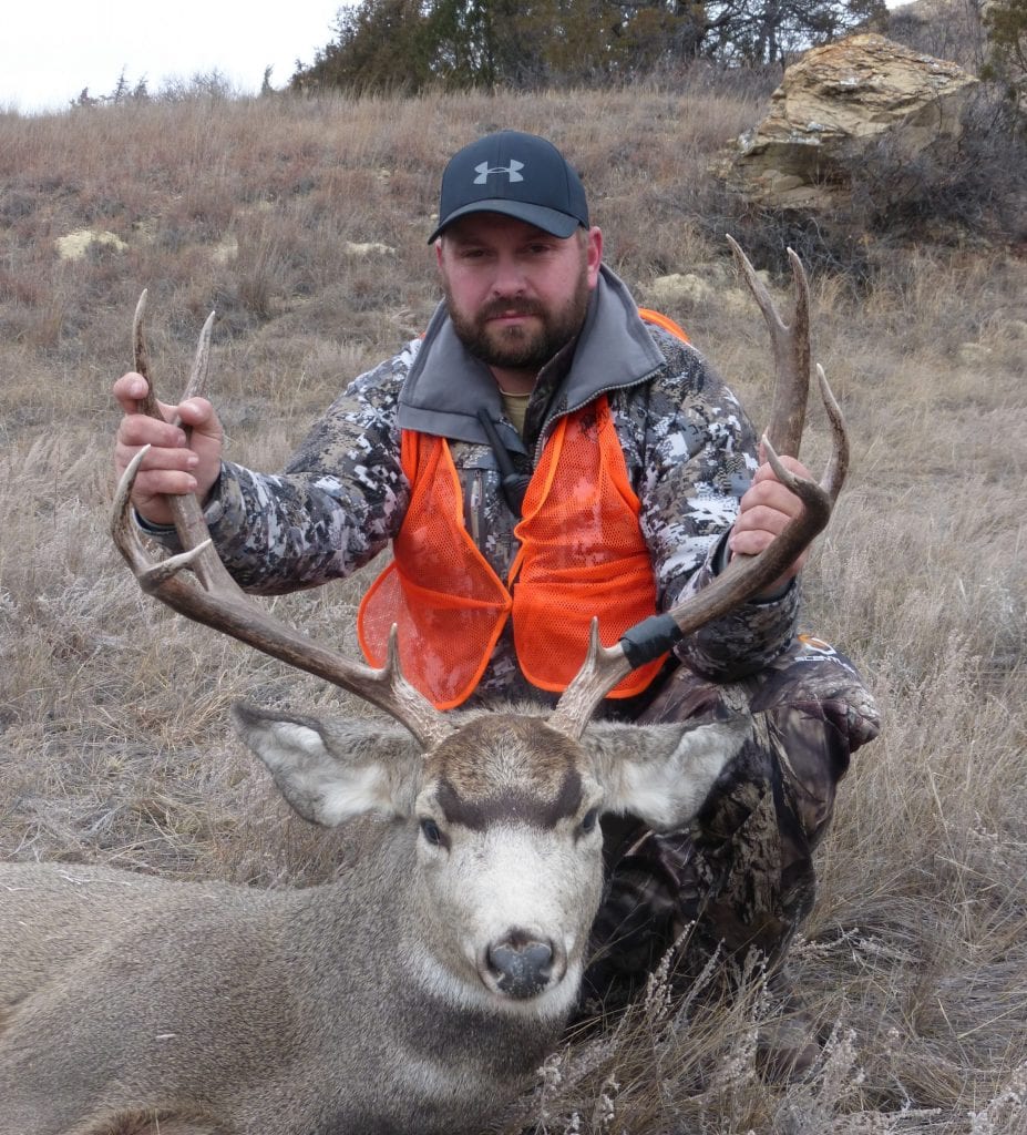 A hunter in camouflage and an orange vest kneels behind a harvested mule deer in a dry, grassy field.