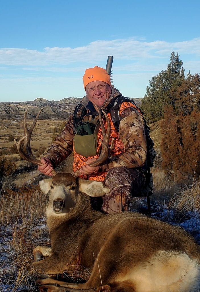 A hunter in camouflage and an orange hat poses with a harvested mule deer in a field with hills in the background.