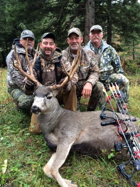 Four hunters pose with a harvested mule deer and a bow in a forest.