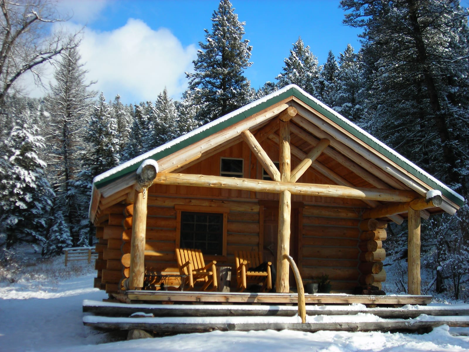A rustic log cabin with a snow-covered roof and porch, surrounded by snow-laden pine trees under a clear blue sky.