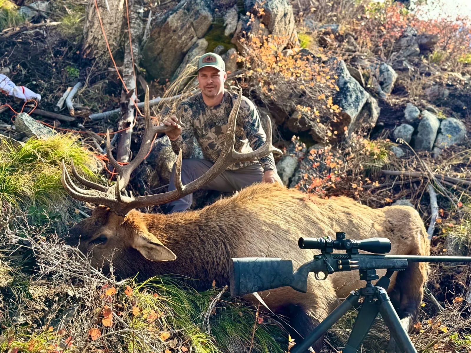 A hunter poses with a harvested elk on a rocky, brushy hillside, with a rifle on a tripod in the foreground.