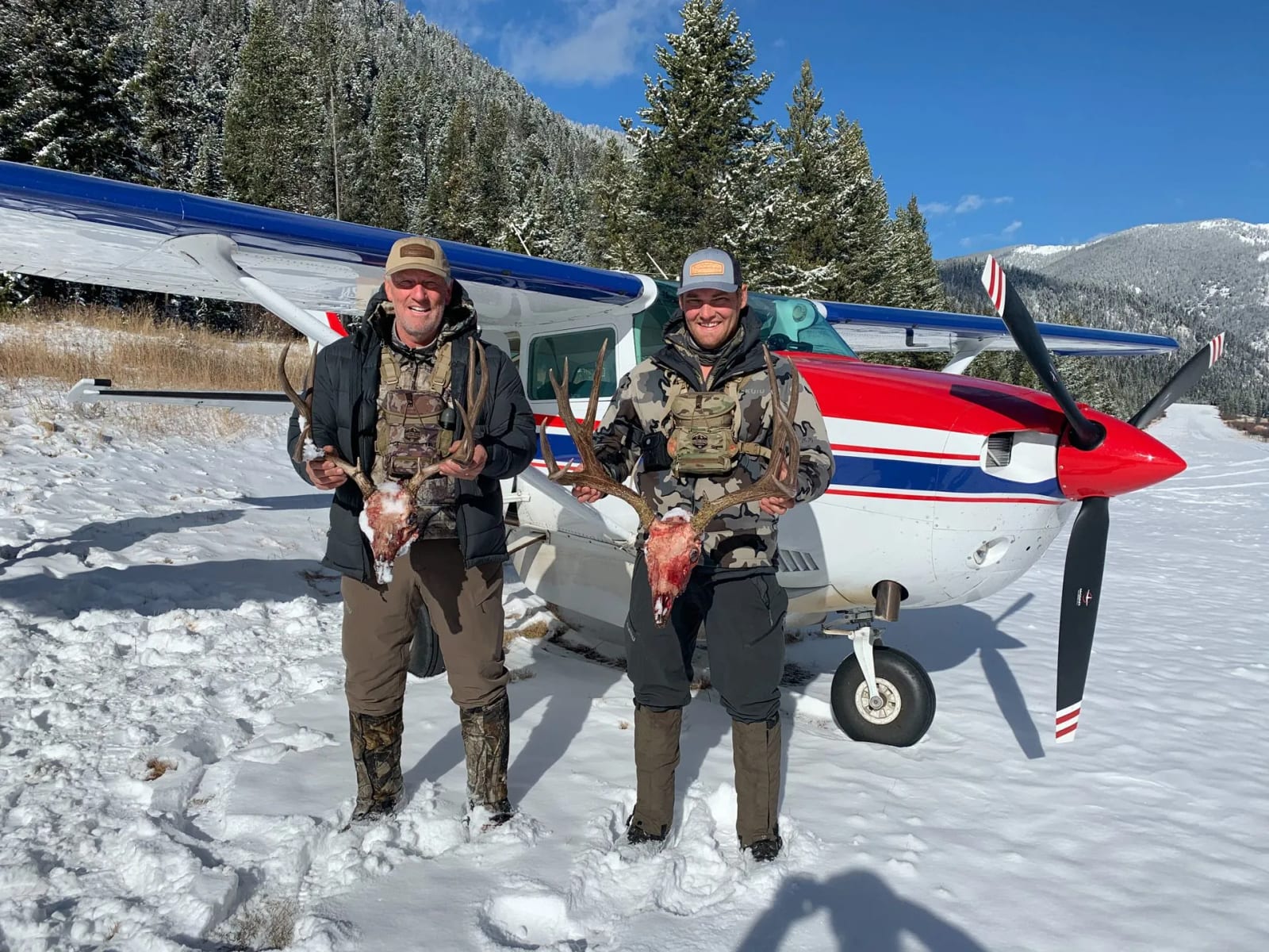 Two hunters pose with harvested mule deer in front of a small plane in a snowy landscape.