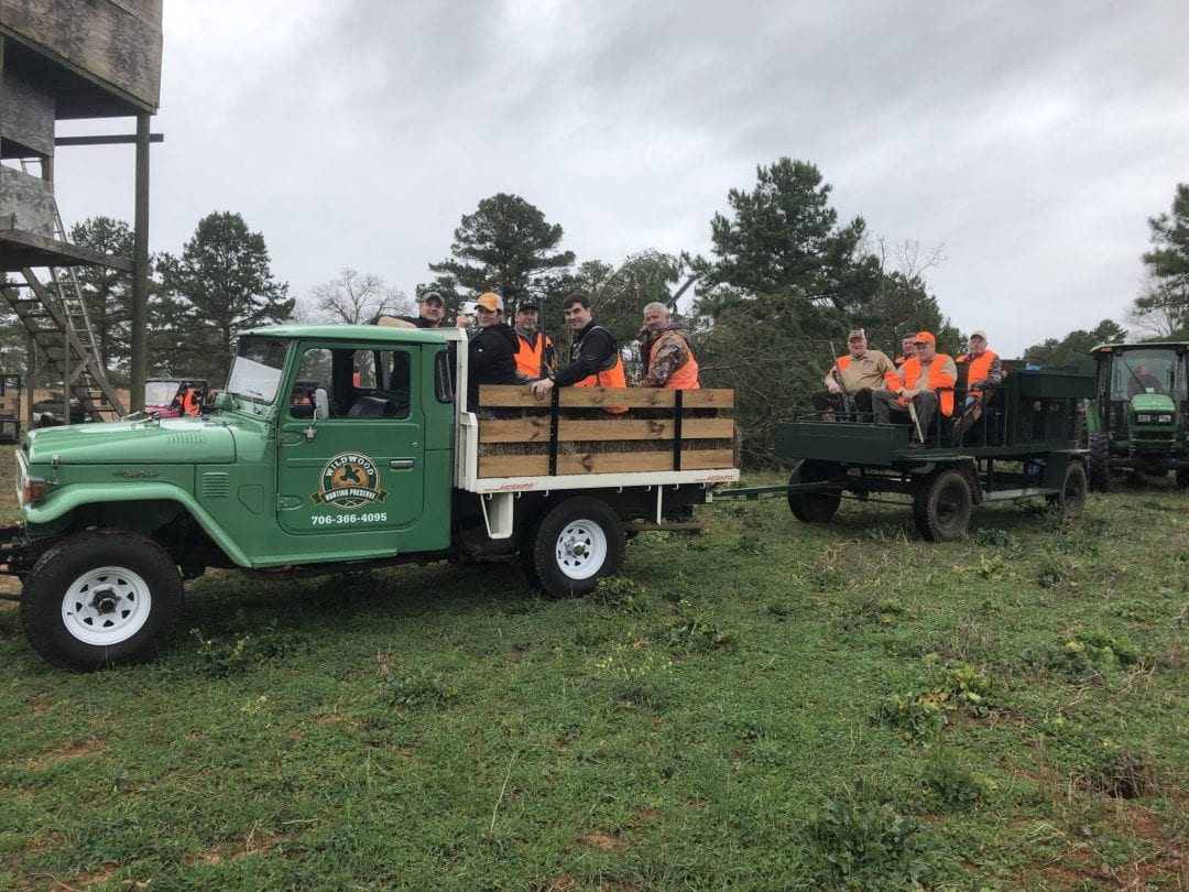 A group of hunters or guides are riding in a green truck and a trailer through a grassy field.