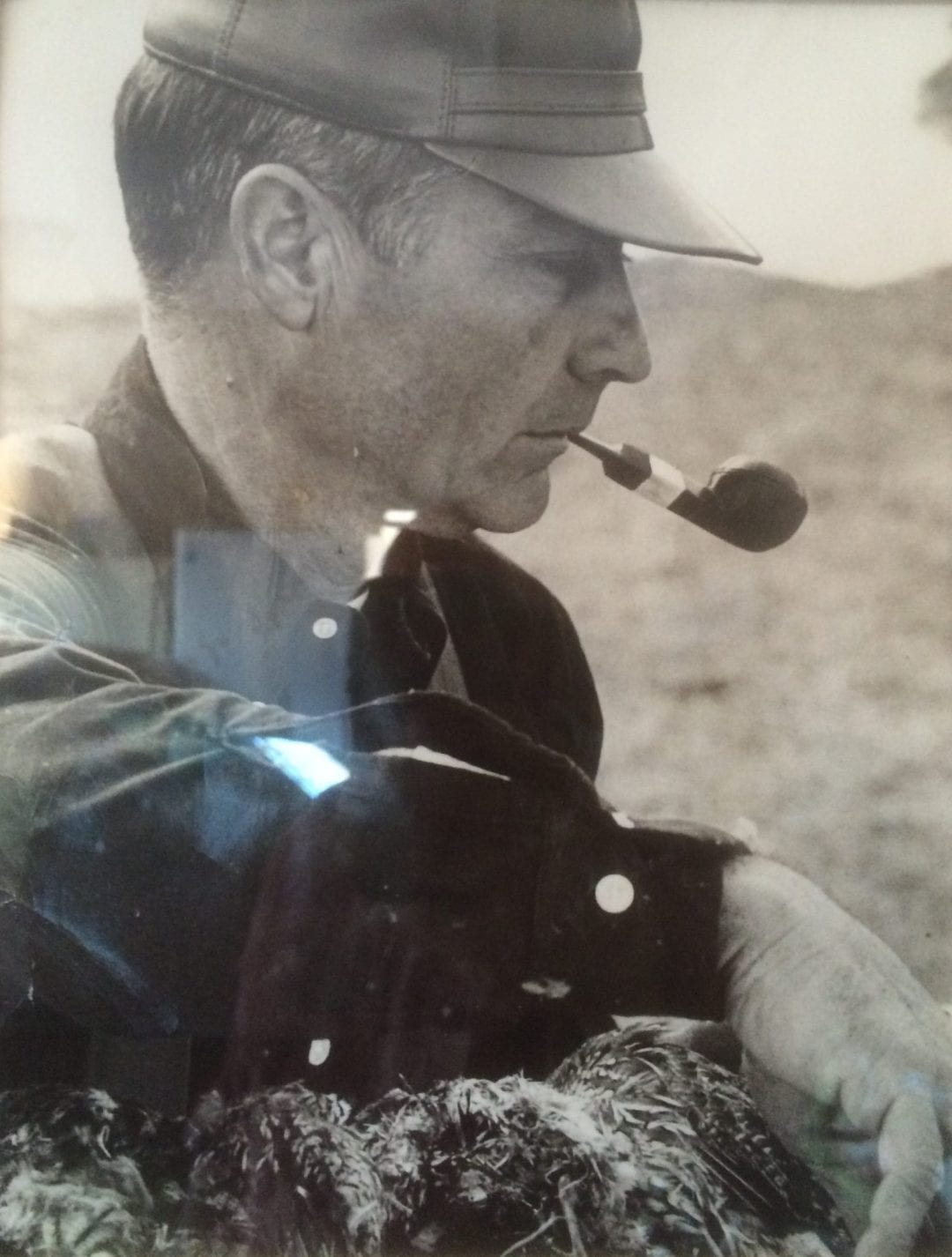 A man in a hat smokes a pipe while holding several harvested quail.