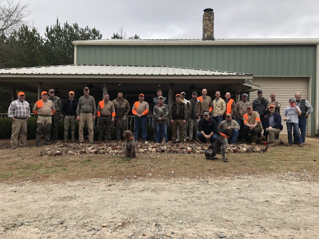 A large group of hunters poses with their harvested northern bobwhite quail and ring-necked pheasants and a hunting dog in front of a lodge building.
