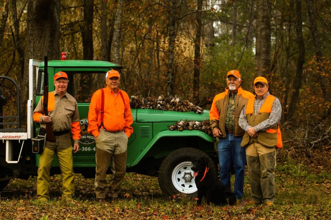 Four hunters and a dog pose with a truck full of harvested northern bobwhite quail in a wooded area.