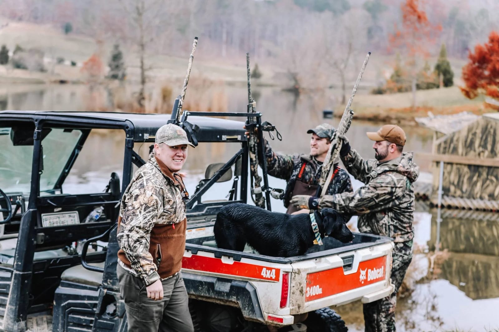 Three hunters in camouflage gear with a black dog on a utility vehicle near a lake, holding rifles, during autumn.