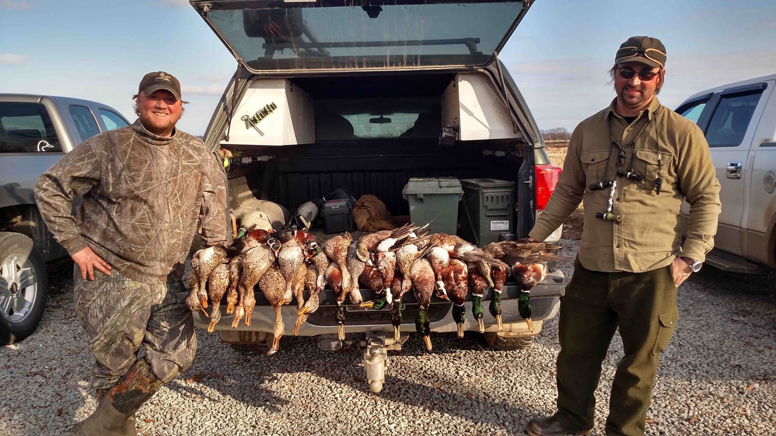 Two hunters pose with their harvested ducks, including mallards and gadwalls, next to a truck.