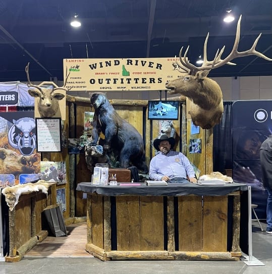 A man sits at a booth for Wind River Outfitters, surrounded by taxidermy mounts of an elk, mule deer, black bear, and wolf.