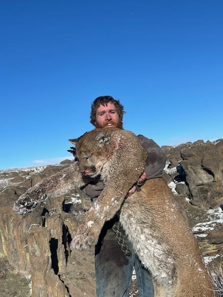 A hunter poses with a harvested mountain lion in a rocky, snowy landscape.