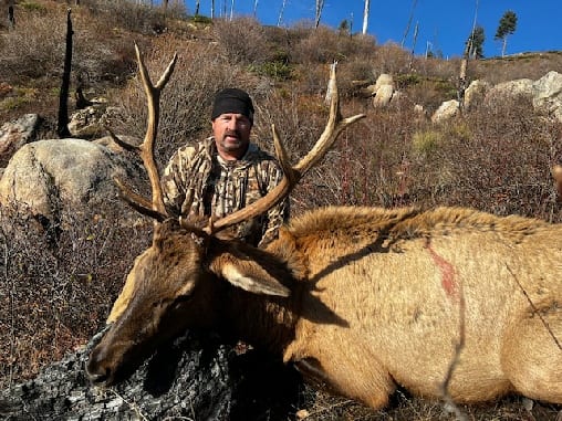 A hunter in camouflage and a black beanie poses with a harvested elk in a rocky, brushy landscape.